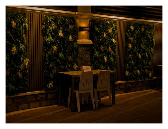 A dark, wooden table with white two chairs neatly arranged at it, and a reserved sign on it. The table is set against a wall, decorated with vertical panels of leaves and wood.