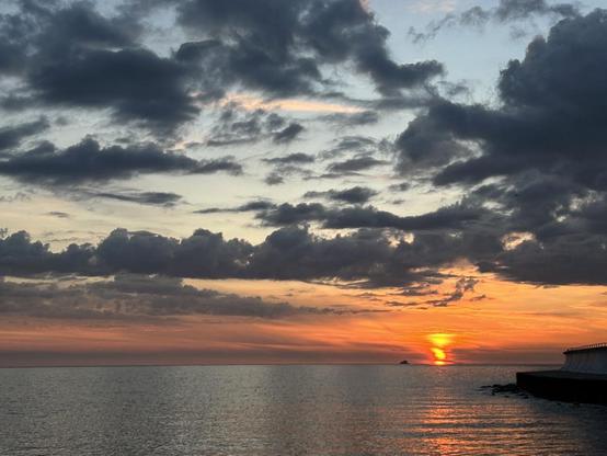 A bright orange-pink-light blue sunrise, partially obscured by dark clouds, over a large body of water with gentle waves reflecting the colors of the sky.