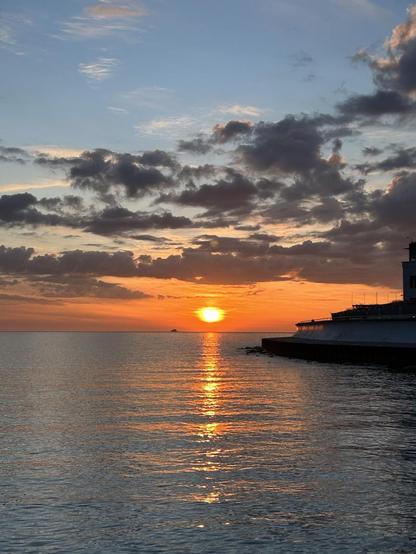 A bright orange-pink-light blue sunrise, partially obscured by dark clouds, over a large body of water with gentle waves reflecting the colors of the sky. To the right is a sea wall and the edge of a building.