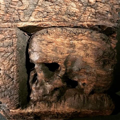 🇩🇪 Das Bild zeigt einen geschnitzten Holzschädel, eingebettet in ein Stück Holz von einem Türrahmen. Der Schädel ist kunstvoll geschnitzt und weist sichtbare Details wie Augenhöhlen, Nasenhöhle und Kieferpartie auf. Das Holz um den Schädel herum ist dunkel und rissig, was auf deutliche Alterung hindeutet. 🇬🇧 The image shows a carved wooden skull embedded in a piece of wood from a doorframe. The skull is intricately carved and features visible details such as the eye sockets, nasal cavity, and jawline. The wood surrounding the skull is dark and cracked, indicating significant aging.