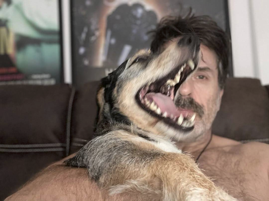 A selfie of Con, shirtless and with his hair a bit mussed, hugging his dog Cooper close to his chest. Con is looking tiredly at the camera, but only half his face is visible, the rest is hidden behind Cooper, whose mouth is wide open, his whole head all blurry as he probably yaps excitedly.