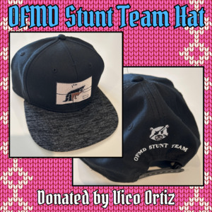 Another item donated by Vico: Vico's Stunt Team Hat. 2 photos show the front and back of a black baseball hat. The front features a patchwork flag with a black cat licking its paw, and the back has the words 'OFMD Stunt Team' written below a picture of a teddy bear's face with an eye patch and a scar on one cheek.