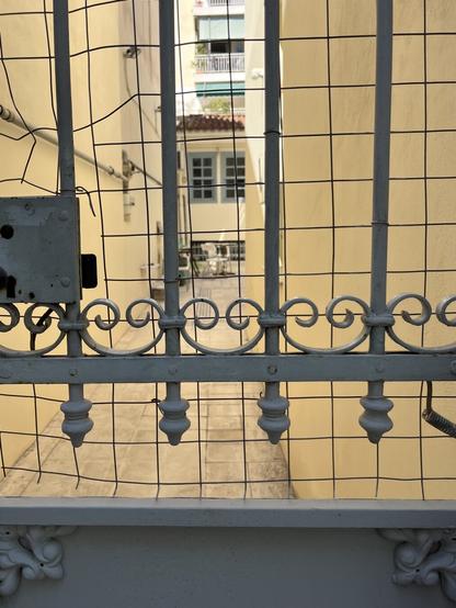 A wrought iron gate painted pale blue leads to a courtyard, cream and pale blue. Behind the low buildings of the courtyard, an apartment block looms.
