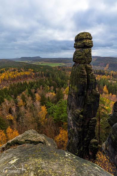 The image shows a striking rock formation in a forest landscape, the Barbarine in Saxon Switzerland. The rock formation is tall and slender, with a layered structure. In the background stretches a forest with trees in various autumn colors. The sky is cloudy.
