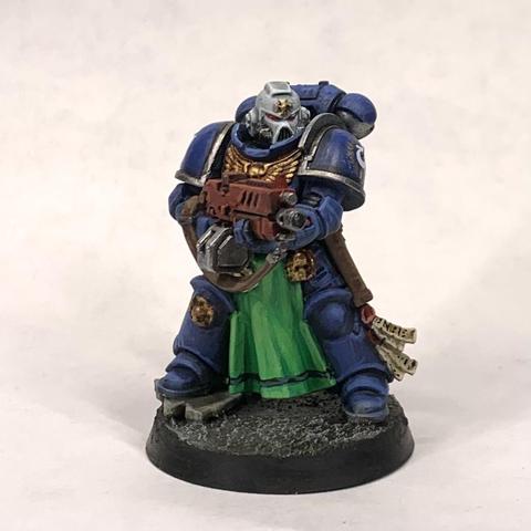 One of squad of blue-armored space marines holding red rifles and in action poses. They stand on tan ground. Their helmets are white except for the squad leader with a red helm with a white stripe. They have green tabards and gold relics hand from their belts.
