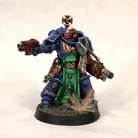 One of a squad of blue-armored space marines holding red rifles and in action poses. They stand on tan ground. Their helmets are white except for the squad leader with a red helm with a white stripe. They have green tabards and gold relics hand from their belts.