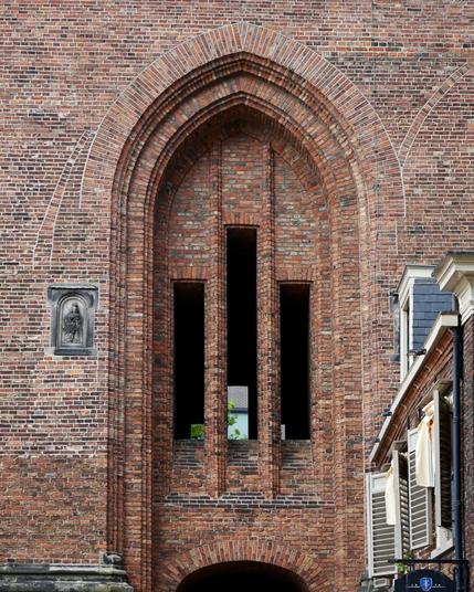 Détail de la façade d’une église en brique gothique montrant une ouverture dont le haut est en ogive. L’intérieur est plein par endroits et percé de trois bandes de vide verticales.
