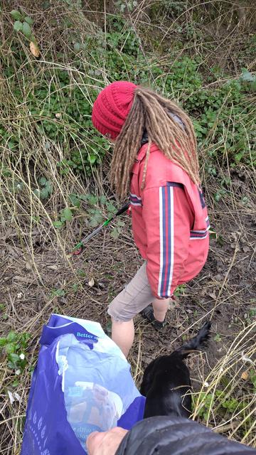 A woman with blond hair in dreadlocks wearing a red knitted hat, red coat and grey shorts in a ditch with a litter picker, in the foreground a hand holding a large blue bag which is being filled with litter.