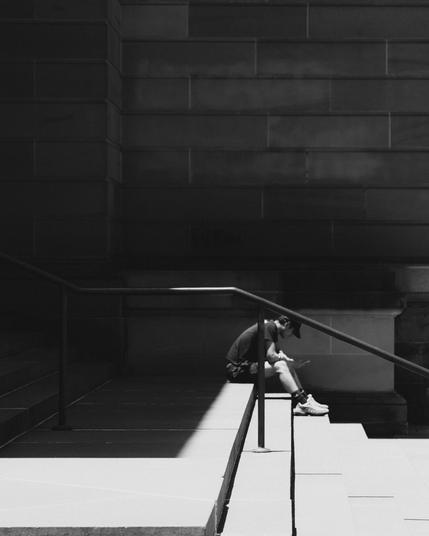 The image depicts a black-and-white scene of a person sitting alone on some steps. The person is leaning forward, possibly looking at a phone or similar device. The lighting creates a stark contrast, with part of the scene in shadow and part illuminated. There's a handrail next to the person, and the setting appears to be urban, possibly near a large building.
