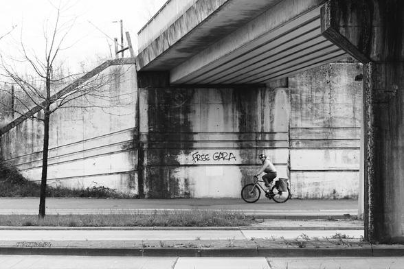 A black and white picture taken under a suburban bridge. On the wall a tag reading «Free Gaza». A cyclist is passing by at that moment and looks in the photographer's direction.