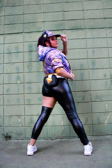 Cosplayer dressed as the character Akali, from the virtual band K/DA, produced by Riot Music/Games. The character wears a cap, top, asymmetrical pants, a holographic purple jacket and accessories.

All the clothes, with the exception of the pants, which are black, are in shades of purple and gold.