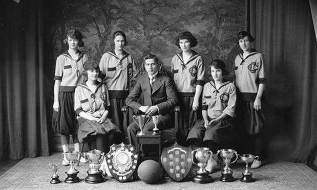 A black-and-white studio photo of six women in early 20th century women's sleeved basketball jerseys and bulky skirts - four standing at the back and two sitting at the front - around a man sitting front and centre. On the floor in front of them are 8 trophy cups, two trophy shield, and a basketball.

Source: City of Edmonton Archives