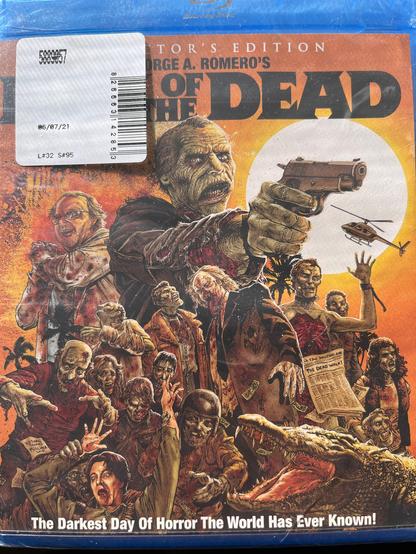 The image shows the cover of a Blu-ray case for "George A. Romero's The Dead." The cover features a central figure, a zombie-like man with a gun, pointing forward. He has a weathered face and is wearing a dark coat. Surrounding him are various zombies in different poses, some reaching out with their hands, others with weapons. The background includes a large moon and a helicopter flying above. The text at the top reads "George A. Romero's The Dead," and at the bottom, it says "The Darkest Day Of Horror The World Has Ever Known!" A barcode and a date "08/07/21" are visible on the top left corner, along with a sticker that includes a barcode and a number "L#32 S#95." The overall color scheme is a mix of orange, red, and brown tones, creating a dramatic and intense atmosphere.