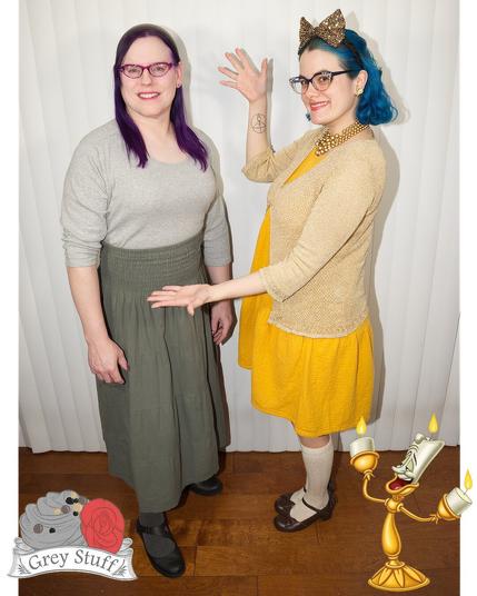 The image features two women standing side-by-side, posing for a photograph. The woman on the left has dark hair and is dressed in a grey top paired with an olive green skirt, complemented by black shoes. Her counterpart on the right boasts blue hair, adorned with cat ears, and wears a yellow dress accompanied by a beige cardigan and white socks.

In the foreground, a small grey stuffed animal resembling a mouse can be seen at their feet, situated to the left of the image. The background is characterized by a plain white wall, while the floor appears to be made of wood. A subtle watermark in the bottom-left corner reads "Grey Stuff" alongside an illustration of a grey mouse.

The overall atmosphere of the image suggests that it was taken during a photoshoot or event, possibly with a theme related to Disney's Beauty and the Beast, given the presence of the cat ears and the mention of "Grey Stuff."