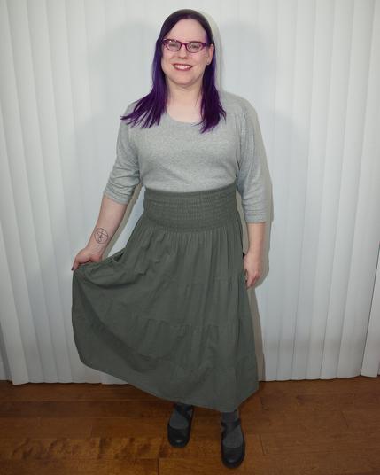 The image features a woman standing against a white wall, her body angled slightly to the left as she faces the camera directly.

She has dark hair that falls just below her shoulders, and is dressed in a gray top paired with an olive green skirt. Her right hand rests on her hip, while her left arm hangs relaxed by her side. The woman's attire is complemented by black shoes, which add a touch of sophistication to her overall appearance.

In the background, a white wall provides a clean and neutral backdrop for the subject, allowing her features and clothing to take center stage.