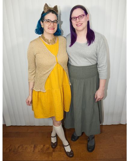 The image features two women standing side-by-side against a white curtain backdrop, their bodies facing the camera directly.

On the left, a woman has short blue hair styled in a bob, adorned with cat ears headbands. She wears a yellow dress complemented by a beige lace cardigan and black Mary Jane shoes. Her outfit is completed with white knee-high socks.

The woman on the right has long purple hair that falls down her back. She sports a gray shirt paired with a green skirt and black flats. The two women stand close together, their arms not visible in the image.

The background consists of a white curtain hanging from the top of the frame to just above the floor, providing a clean and simple setting for the subjects.