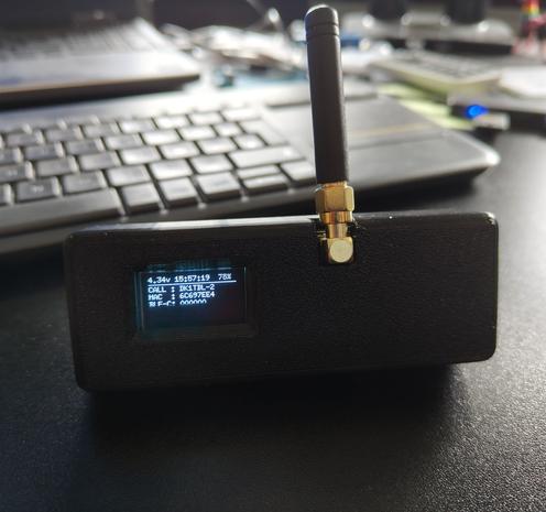 Auf einem Schreibtisch steht ein T-Beam-Device in einem schwarzen 3D-Druck-Gehäuse. Oben befindet sich eine Antenne. Links sieht man ein kleines Display.