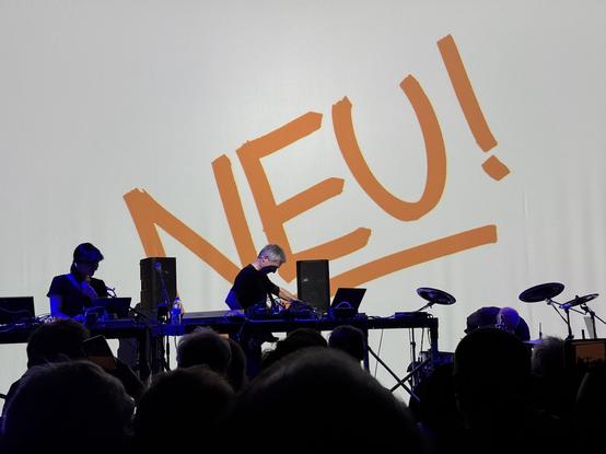 Michael Rother onstage at the Grey Area in San Francisco, March 24, 2025. Vittoria Maccabruni is on the left, on keyboards and vocals, while Hans Lempe on drums is in the lower right. The cover of the debut Neu! album, with the band name in red against a white background, is projected behind him.