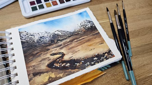 Open sketchbook on a desk showing a landscape of a dried river bed vanishing into distance with stones in the middle. Dried fields of yellow grass and sand around it and snowy topped mountains in the back. White clouds are framing the mountain tops showing only hints of a blue sky.
Next to the sketchbook are a few brushes... ready to paint more landscapes just as a reson for the mind to wander to beautiful places that are not here and now.