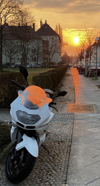 Das Bild zeigt einen weißen Motorroller, der an einem gepflasterten Bürgersteig geparkt ist. Der Windschutz ist beschlagen und leuchtet orangefarben. Im Hintergrund ist ein Sonnenuntergang zu sehen, der den Himmel in warme Orangetöne taucht. Die Sonne steht tief am Horizont und reflektiert sich auf den Bürgersteig. Links im Bild sind einige Bäume und ein gepflegter Heckenstreifen zu sehen, während rechts mehrere geparkte Autos und ein weiterer Bürgersteig zu erkennen sind. Im Hintergrund sind mehrere Gebäude mit Dächern und Fenstern zu sehen, die typisch für eine städtische Umgebung sind. Die Szene wirkt ruhig und friedlich, mit dem Motorrad als zentrales Element im Vordergrund.

Bereitgestellt von @altbot, privat und lokal generiert mit Ovis2-8B

🌱 Energieverbrauch: 0.233 Wh