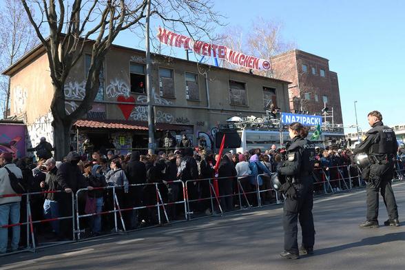 Das Foto zeigt demonstrierende Menschen hinter Hamburger Gittern, am About Blank, auf dem ein Transparent mit dem Text "Antifa. Weiter. Machen" hängt. Davor steht der "Adenauer SRP+" mit Laufschrift auf dem Dach: "Nazi go Home / Fuck Nazipack!" usw.