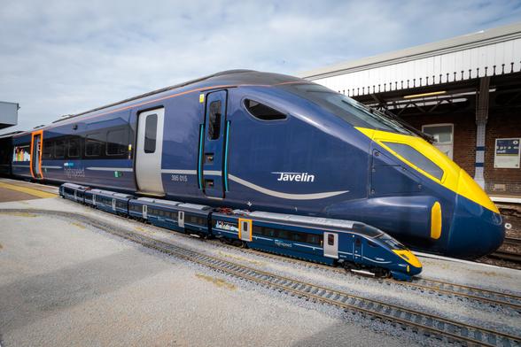 The image shows a large, modern passenger train positioned on railway tracks beside a miniature model of the same train. The full-sized train is sleek, with a dark blue body and vibrant yellow accents on the front. It is labelled "Javelin" and carries the number "395 015" on its side. The windows are tinted, and there are detailed markings in light blue and white. The model train on the gravel beside it mirrors the full-sized train’s design, including the colours and labelling, but is much smaller in scale. The setting is an open-air station with part of a white-roofed platform visible in the background under a cloudy sky.

Photo by Southeastern