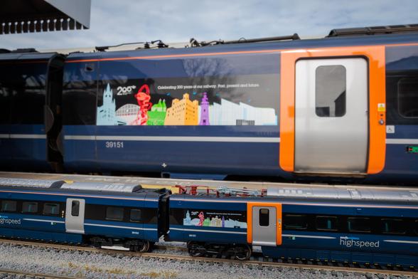 The image shows a close-up of two trains on parallel tracks. In the background, a full-sized train is partially visible, featuring a large colourful graphic with silhouettes of buildings and a spiral element. The text on the graphic celebrates a 200-year railway anniversary. The train is painted in dark blue with orange accents and has a visible door with a window. In the foreground, a model train sits on a gravel-like track. This model replicates the larger train with similar blue and orange colours, maintaining the same graphic and text on a smaller scale. The image captures both trains in a side view, highlighting their design and anniversary theme.

Photo by Southeastern