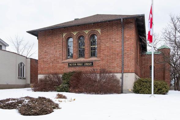 A brick building with arched windows and a sign reading Weston Public Library is surrounded by snow-covered ground and bushes. A Canadian flag is on a pole to the right. Leafless trees are in the background.