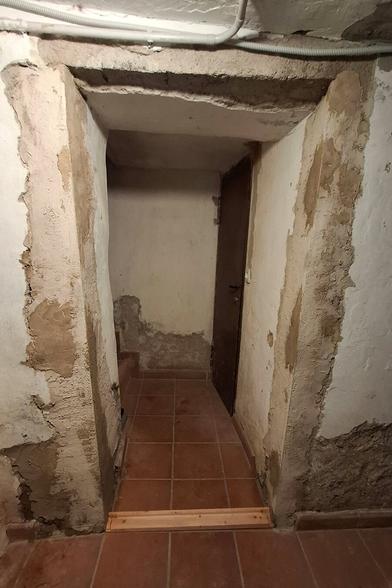The scene shows a narrow hallway leading to a door at the end. The walls have a rough, partially plastered finish. The door frame and threshold have been adapted with wooden trim, which aligns with a pine wood pieces for a precise fit.
The floor is tiled, and there is a nwooden threshold strip at the entrance, not yet fixed and with two screws on the ground. The lighting and overall condition suggest an old cellar space undergoing restoration. The walls still show some signs of moisture and unfinished areas, indicating ongoing work.