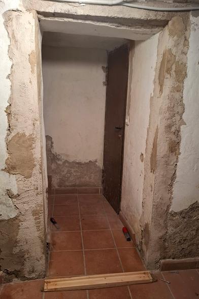 The scene shows a narrow hallway leading to a door at the end. The walls have a rough, partially plastered finish. The door frame and threshold have been adapted with wooden trim, which aligns with a pine wood pieces for a precise fit.
The floor is tiled, and there is a newly installed wooden threshold strip at the entrance, secured with screws. The lighting and overall condition suggest an old cellar space undergoing restoration. The walls still show some signs of moisture and unfinished areas, indicating ongoing work.