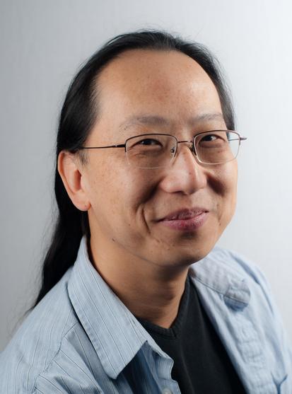 Head-and-shoulders color photo of Curtis C. Chen, a man of East Asian descent. He has medium-dark warm-toned skin and wears his long black hair brushed back, showing power alleys. He wears rectangular wire-rimmed glasses and a light blue button-up over a dark T-shirt and. He poses in a three-quarters view against a light, neutral background. He regards the camera with a mischievous smile, like something’s cracking him up. Photo credit: Folly Blaine
