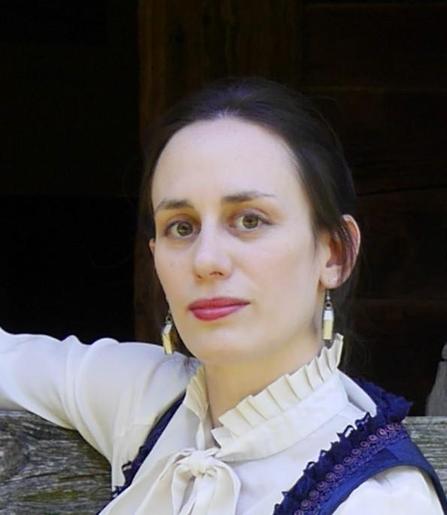 Head-and shoulders color photo of Sonia Focke, a fair-skinned woman with dark eyes and long, dark hair worn swept back. She wears a ruffled dark blue vest over a white blouse with a high, pleated collar and a bow; long, dangling white earrings; and light makeup with a dark pink lipstick. She poses against a dark background, one arm raised as if resting on something, and looks into the camera in a three-quarters view. Chin lifted, she gives us a small and knowing smile.