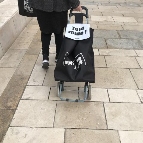 Dans la rue une personne tire un chariot de course noir et blanc où est écrit : " Tout roule ! "