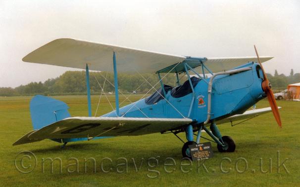 Side view of a pale blue biplane with white wings parked on grass facing to the right.
The plane has 2 cockpits, covered with blue rubber covers to protect them from the elements.
There is a red, yellow, and white triangle with white "Spartan Arrow" titles on top and below, under the front cockpit.
Grass fills most of the frame, with trees in the distance, with a couple of other light aircraft parked in front, under grey skies.