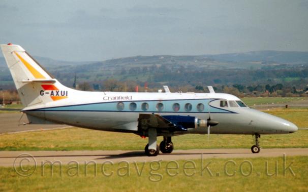 Side view of a white and grey, twin propellor-engined airliner taxiing from left to right.
The plane has white upper surfaces and grey lower surfaces, with a pale blue stripe running along the body, and black "Cranfield" titles on the upper rear fuselage.
There is a diagonal orange stripe on the tail, with the black registration "G-AXUI" passing through the base.
The wing-mounted engine pods are dark blue, almost purple.
Green grass fills the foreground. With more grass in the background leading up to trees in the distance, with tree-covered rolling hills beyond that.
Grey sky fills the rest of the frame.