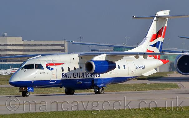 Side view of a white, high-winged, twin engined jet airliner taxiing from right to left.
The plane has a deep blue belly and engine pods slung under the wings, and blue "British airways" titles on the lower forward fuselage.
There is a twisted ribbon, blue on the inside, red on the outside, on the upper forward fuselage, covering part of the forward cabin door.
There are wavy red-blue-red stripes, separated by white, on the white tail.
The registration "OY-NCM" is on the rear fuselage, in blue.
There are small "Operated by Sun-Air" titles under the cockpit windows.
Grass lines the runway in the foreground, while a grey terminal building stretches across the frame, varying in height, taller on the left, barely reaching over the top of the plane on the right.
Blue-grey sky fills the rest of the frame.
