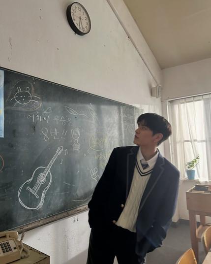 Seungmin stands in a classroom, looking sideways at a blackboard behind him. A guitar and a couple of cats(?) are drawn on the blackboard. "Anyway, class 8 will win!!" is written in Korean