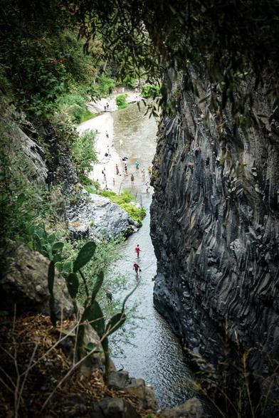 The image depicts a scenic natural landscape featuring a narrow river flowing through a rocky gorge. The river is flanked by steep, dark volcanic rock formations on both sides, with a sandy beach area visible in the background where several people are seen enjoying the water and the surroundings. The foreground includes lush green vegetation, such as cacti and shrubs, framing the view. The water appears calm, reflecting the surrounding rocks and vegetation. The scene is framed by hanging branches, adding depth and a sense of enclosure to the view. The overall atmosphere is one of natural beauty and tranquility, with people engaging in recreational activities.

Provided by @altbot, generated privately and locally using Ovis2-8B