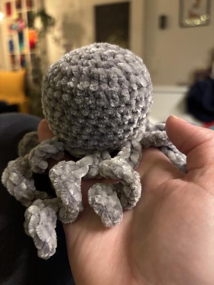 Auf einer Hand sitzt ein grauer gehäkelter Oktopus aus sehr flauschigem Garn.
Die Tentakeln sind in sich gedreht.
Im Hintergrund ist verschwommen ein Wohnraum zu erkennen.