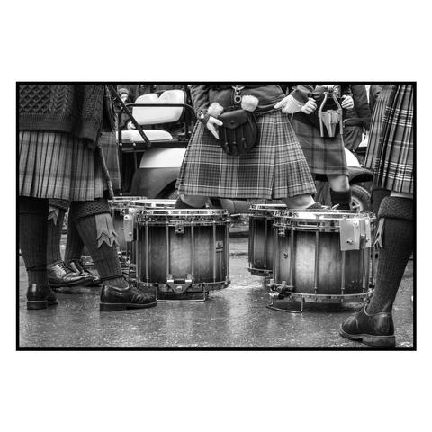 (English follows) Photographie noir et blanc, format paysage, ratio 3x2. L'image est prise à hauteur de taille, montrant le bas du corps de trois personnes d'un ensemble de musique, et leurs grosses caisses. Les personnes sont vêtues d'un kilt irlandais : kilt, la bourse portée à la ceinture (sporran) et les hautes chaussettes de laine avec leurs flashs (kilt hose). 
Black and white photograph, landscape format, 3x2 ratio. The image is taken from waist height, showing the lower body of three people in a music ensemble, and their bass drums. The people are dressed in an Irish kilt: kilt, purse worn on the belt (sporran) and high woollen socks with their flashers (kilt hose).