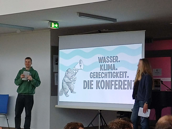 zwei Moderator*innen stehen auf einer Bühne vor einer Projektion mit Schrifzug: "Wasser. Klima. Gerechtigkeit. Die Konferenz", daneben eine Graphik eines Bibers mit Megaphon
