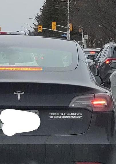 a Tesla with a bumper sticker 'I bought this before we knew Elon was crazy'.  traffic lights are visible in the background and other cars are stopped at the light