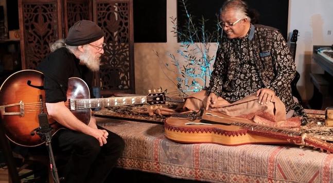 Steve kimock in a black t-shirt and holding a large Gibson guitar, looking over at Debashish Bhattacharya, wearing a kurta with intricate designs looks back at him smiling. His Indian slide guitar is at his feet.