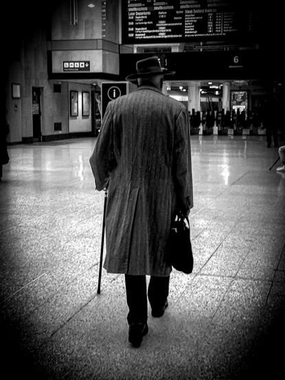 Black and white. An elderly man in a knee-length overcoat, wearing a black fedora hat and carrying a black shopping bag, uses a walking stick as he walks away from the viewer towards platform barriers in a train station.