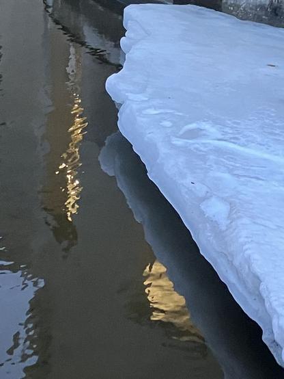 Water of the river flows under a previously floating shelf of ice that now hovers in the air, supported by the building it is against. Some lights reflect on the water surface.