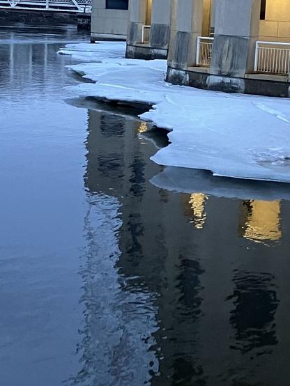 Further away photo of previous scene. Water of the river flows under a previously floating shelf of ice that now hovers in the air, supported by the building it is against. Some lights reflect on the water surface.