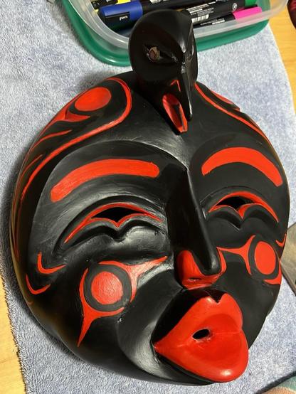 Hand-carved and painted mask in the style of Northwest Coast Indigenous art, featuring a human face in black with red highlights and a small, stylized Raven figure perched on top. The mask has abstract shapes around the eyes and mouth.