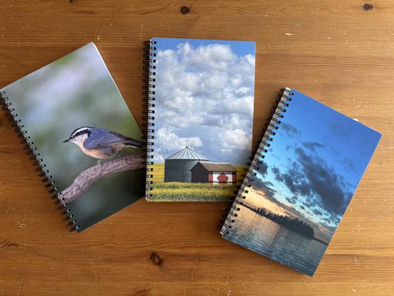 Three coil-bound journals displayed on a table surface. The covers include: a red-breasted nuthatch on a branch; a grain shed and small barn with the Canada flag painted on it, in a canola field; and a sunset behind an island in the middle of the iconic Astotin Lake in Alberta.