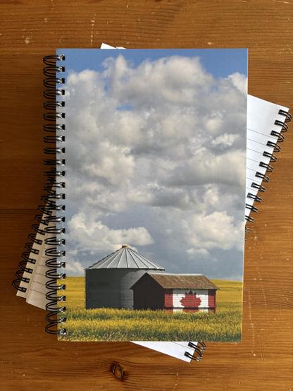 Coil-bound journal stacked on top of others. Cover shows a grain shed and small barn with the Canada flag painted on it, in a canola field.