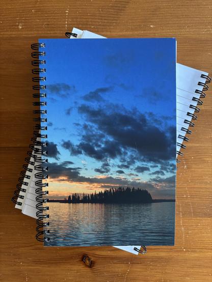 Coil-bound journal stacked on top of others. Cover shows a sunset behind an island in the middle of the iconic Astotin Lake in Alberta.